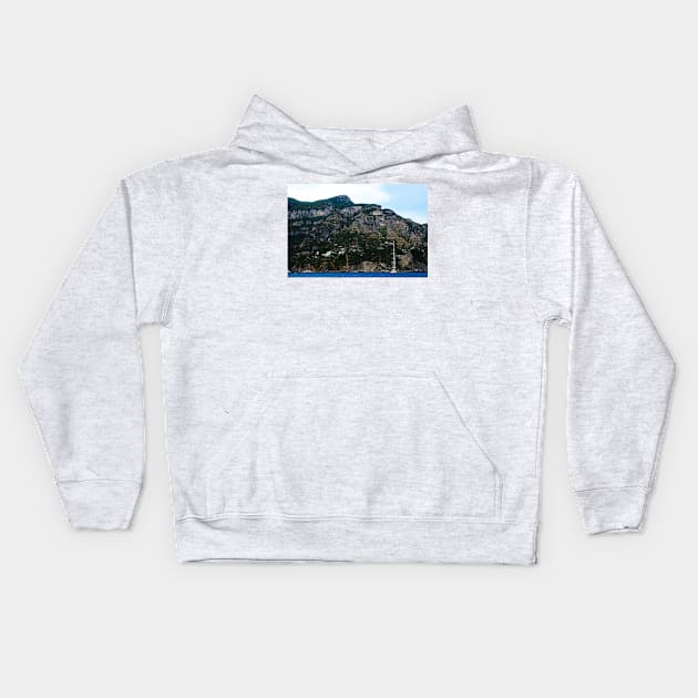 View in Amalfi coast at a huge mountain with rocky parts, greenery and buildings with a white boat underneath Kids Hoodie by KristinaDrozd